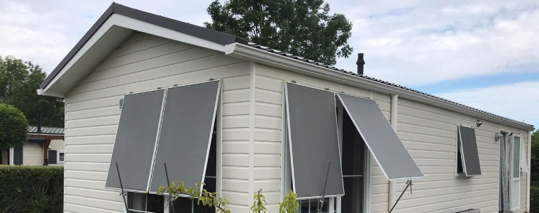 Een chalet met witte gevelbekleding, grijze zonneschermen en een zadeldak, omgeven door groene struiken en bomen.