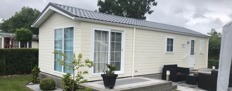 Een modern chalet met witte gevelbekleding, grote ramen en een grijs zadeldak, gelegen naast een betegeld terras en een goed onderhouden tuin.